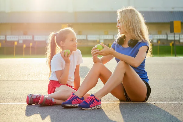 Junge und schöne blonde Frau zusammen mit einem kleinen Mädchen in Sportbekleidung auf dem Asphalt sitzen und essen einen grünen Apfel im Stadion. aktive Mutter und Tochter joggen im Sommer morgens — Stockfoto