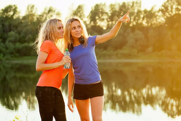 Active family at outdoor. two flexibility curly blonde beautiful woman twin sisters in stylish sportswear have a rest and photographing selfie phone in the stadium lake nice sunset background park — Stock Photo, Image