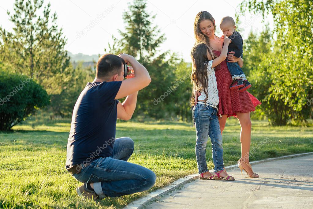 happy family is resting in the park in summer . beautiful mother , handsome father with a little baby boy and cute daughter photographed on camera . family photo shoot male photographer