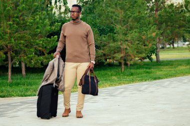 Genç ve yakışıklı model Afro-Amerikan şık takım elbiseli, yaz parkında Latin Latin iş adamı zengin bir turist havaalanına bavuluyla konferansa gidiyor.