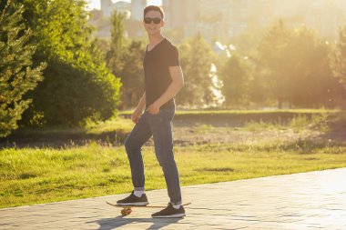 Güneş gözlüklü, kot pantolonlu ve yaz parkında kaykay yapan havalı bir adam.