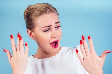 close-up portrait of nervous unhappy young blonde woman looking at a broken fingernail and crying . red long nails manicure broken nail blue background in studio clipart