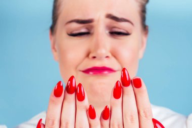 close-up portrait of nervous unhappy young blonde woman looking at a broken fingernail and crying . red long nails manicure broken nail blue background in studio clipart