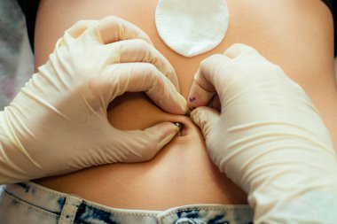 Ustanın yakın plan lastik eldiveni, karnında bandaj ve pamukla genç bir kadının karnını delmeye hazırlanıyor. göbek halka piercing işlemini başlat