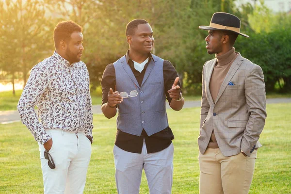 Um grupo de três homens negros em ternos elegantes em um parque de verão. Afro-americanos amigos hispânico empresário comunicando ao ar livre — Fotografia de Stock