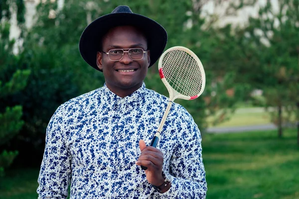 a young and handsome stylish model afro- american man in a stylish suit and black hat holding a tennis racket badminton in a summer park. latino american hispanic businessman black guy resting