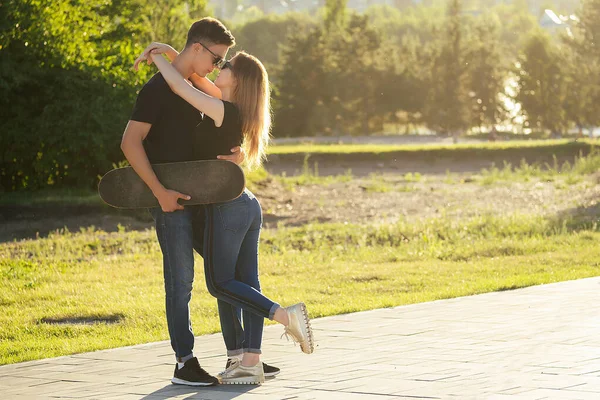 Verliefd stel knuffelend in het zomerpark. de jonge kerel houdt een skateboard vast en kust zijn lieve mooie vriendin close-up — Stockfoto