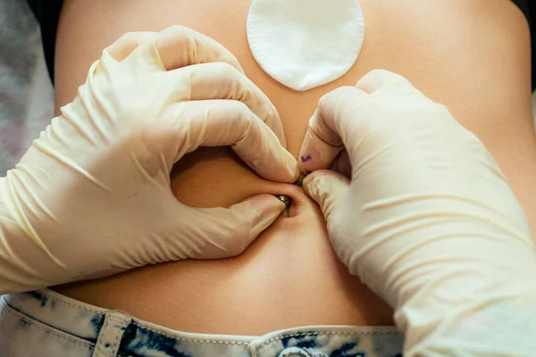 hand in a rubber glove close-up of master prepares to pierce the navel by belly of a young woman with a bandage and cotton on her stomach. process navel ring piercing