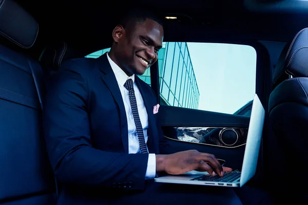 Handsome successful rich african american business men chief in a stylish black suit and tie sitting in a luxury car and works with laptop. concept of luck and currency market