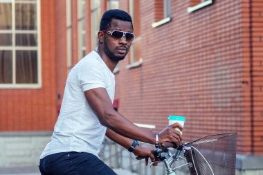 Havalı ve genç Afrikalı Amerikalı bir adam elinde kahve bardağıyla yaz parkında bisikletin üstünde oturuyor.