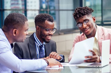 Moda sektöründe iş ortağı üç şık Afro-Amerikan iş ortağından oluşan bir grup açık havada bir kafede dizüstü bilgisayarla masa başı menkul kıymetler imzalama toplantısı yapıyor.
