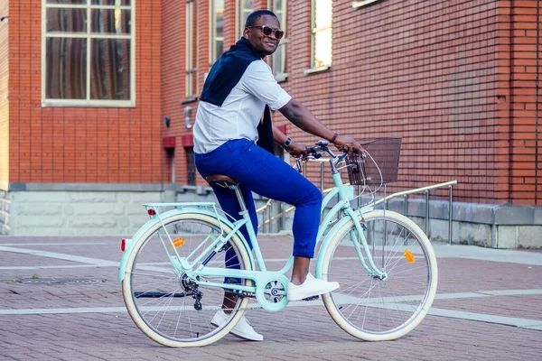 Guapo hombre afroamericano elegante bien vestido con gafas de pie con una bicicleta azul rojo ladrillo edificio de fondo. concepto de transporte ecológico deportivo — Foto de Stock