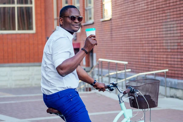 Hombre afroamericano elegante y joven sosteniendo un vaso con café sentado en una bicicleta en el parque de verano — Foto de Stock