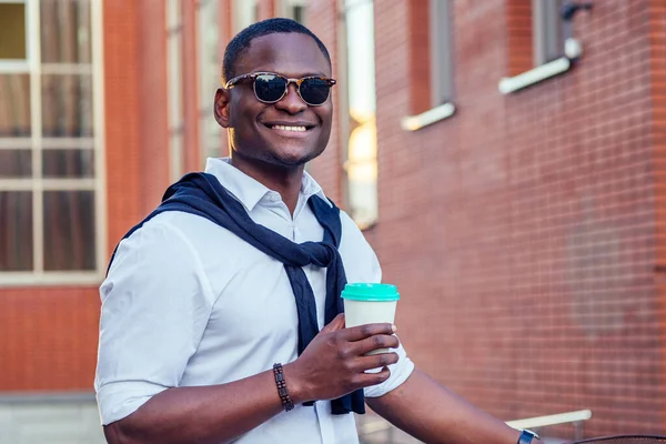 Κομψό και νεαρός αφρικανός φοιτητής Αμερικανός άνθρωπος κρατώντας γυαλί με καφέ το καλοκαίρι στο πάρκο κόκκινο τούβλο κτίριο φόντο — Φωτογραφία Αρχείου