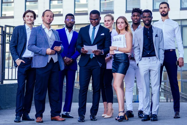 Um grupo de sete empresários bem sucedidos empresária e seus parceiros masculinos em ternos elegantes de pé e olhando orgulhosamente ao ar livre. o trabalho em equipa e o conceito de empresa multiétnica — Fotografia de Stock