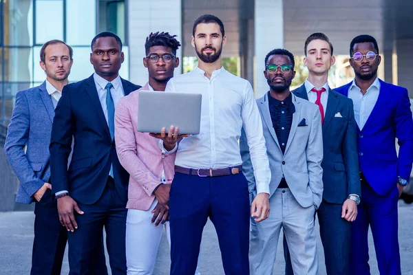 Un grupo de siete hombres de negocios de éxito en trajes elegantes de pie y buscando orgullosamente al aire libre. el trabajo en equipo y el concepto de empresa multiétnica —  Fotos de Stock