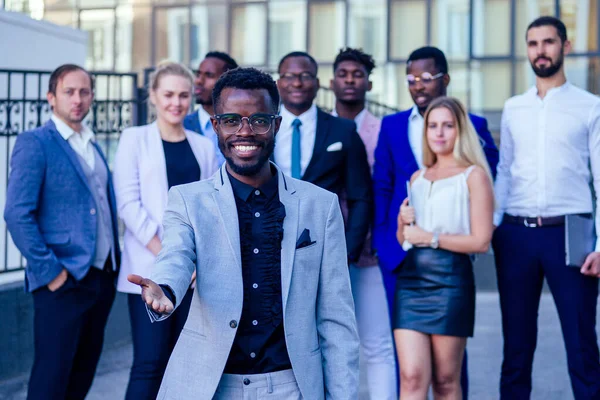 Framgångsrik och stilig afroamerikansk man i en snygg kostym ger en hand för en handskakning tecken välkommen inför en grupp en hel del människor multiracial multietniskt företag — Stockfoto