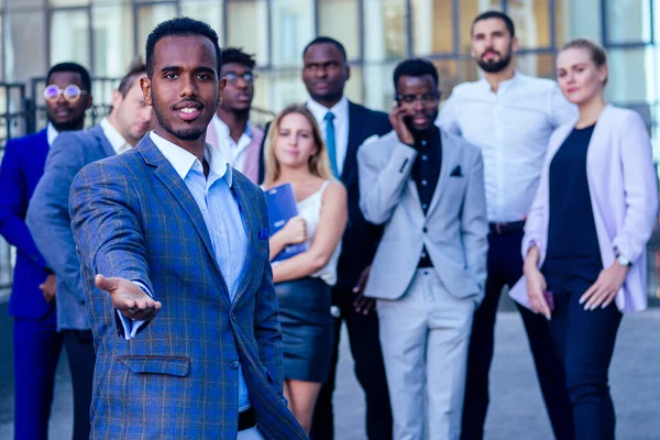 successful and handsome African American man in a stylish business suit giving a hand for a handshake sign welcome ahead of a group a lot of people multiracial multiethnic company