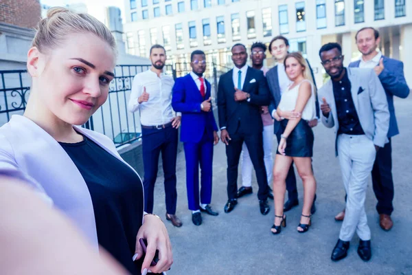 Επιτυχημένη ομάδα εννέα πολυεθνικών επιχειρηματιών σε ένα κοστούμι επιχειρήσεων κοιτάζοντας το τηλέφωνο και παίρνει φωτογραφίες selfie στέκεται εταιρεία των ανθρώπων στο δρόμο . — Φωτογραφία Αρχείου
