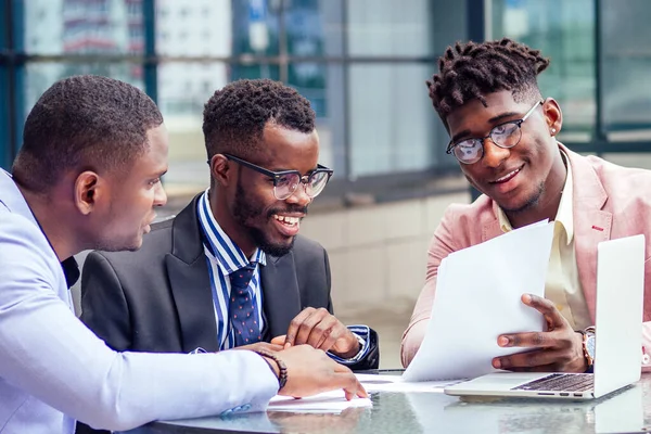 Un grupo de tres empresarios afroamericanos con estilo se asocia con empresarios en trajes de negocios de moda que se reúnen sentados a la mesa y firman los valores con la computadora portátil en un café de verano al aire libre —  Fotos de Stock