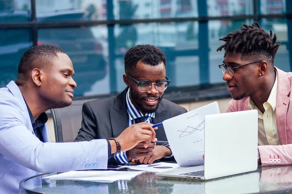 Μια ομάδα από τρεις κομψούς Αφροαμερικανούς επιχειρηματίες φίλους επιχειρηματίες με κοστούμια μόδας συνάντηση στο τραπέζι με φορητό υπολογιστή σε ένα καλοκαιρινό καφέ σε εξωτερικούς χώρους — Φωτογραφία Αρχείου