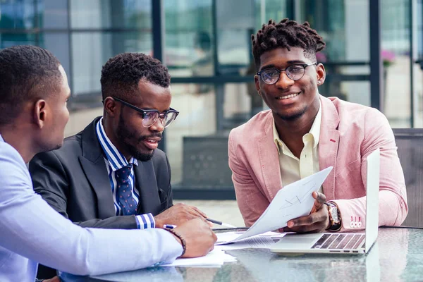 Un grupo de tres empresarios afroamericanos con estilo en trajes de negocios de moda que trabajan sentados en la mesa con el ordenador portátil en un café de verano al aire libre —  Fotos de Stock