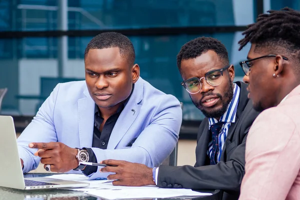 Un grupo de tres hombres afroamericanos con estilo empresarios en trajes de negocios de moda que trabajan sentados en la mesa con el ordenador portátil en un café de verano al aire libre —  Fotos de Stock