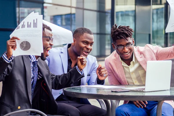 A group of their three successful African American businessmen in a stylish suit sit at the table and work with a laptop and papers skyscraper window background. teamwork and success concept — Stock Photo, Image