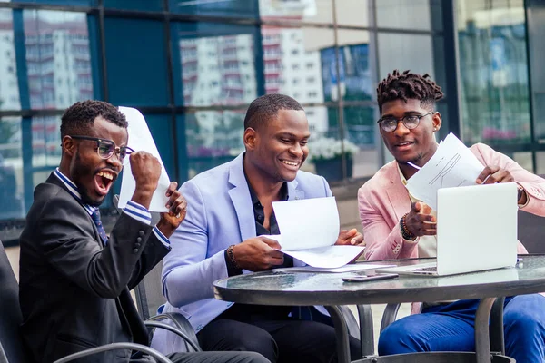 Un grupo de sus tres exitosos hombres de negocios afroamericanos en un elegante traje se sientan a la mesa y trabajan con una conferencia en línea para computadoras portátiles y vomitan papeles. Trabajo en equipo y concepto de plazo —  Fotos de Stock