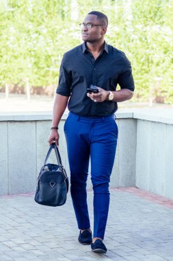 şık ve yakışıklı Afrikalı iş adamı mavi pantolonlu ve siyah tişörtlü bir adam sokakta yürürken çantayı tutuyor.