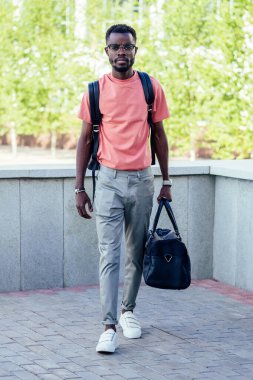 Gri pantolonlu ve pembe tişörtlü yakışıklı Afrikalı öğrenci sokakta yürürken çantasını tutuyor.