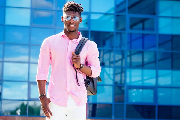 Bonito moderno afro-americanos rastaman macho dreadlocks cabelo com óculos em roupas elegantes colorido t-shirt com uma mochila sobre os ombros stand fundo das janelas azuis da universidade — Fotografia de Stock