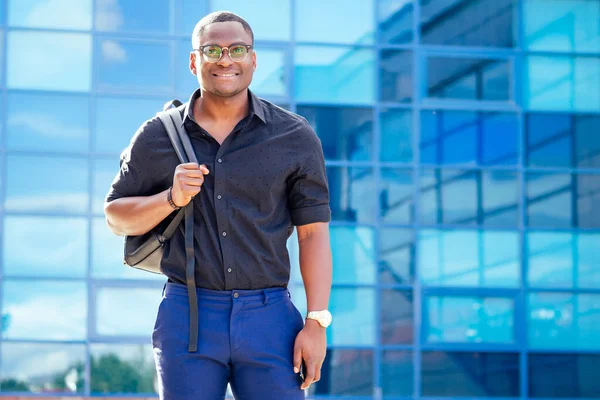 handsome trendy African American university lecturer male with glasses in stylish clothes black shirt with a backpack on the shoulders stand background of the blue windows
