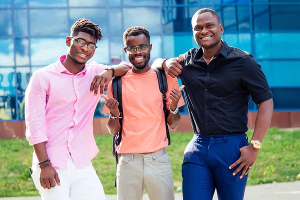 En grupp av tre afroamerikaner manliga vänner i snygga kläder står mot bakgrund av de blå fönstren på universitetet — Stockfoto