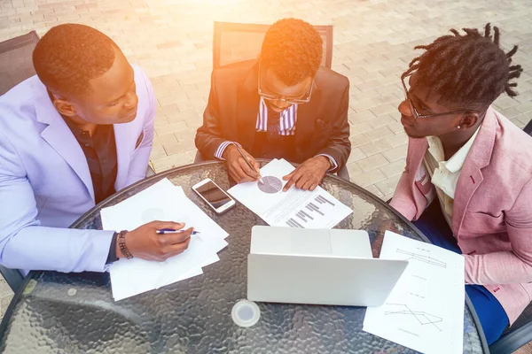 Grupo de três negros afro-americanos em terno de negócios elegante, relógio de pulso caro e gravata sentar na mesa redonda com laptop, títulos e contratos. conceito de negócio bem sucedido e bom negócio — Fotografia de Stock