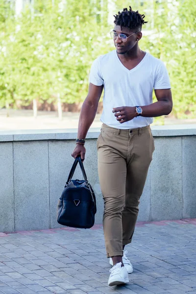 Homem afro-americano elegante e bonito com dreadlocks frescos nas calças marrons e um olhar branco da forma da T-shirt que prende o saco que anda na rua — Fotografia de Stock