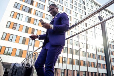 İyi giyimli ve başarılı bir Afrikalı Amerikalı iş adamı havaalanına giderken tekerlekli bavuluyla telefon ekranına bakıyor.