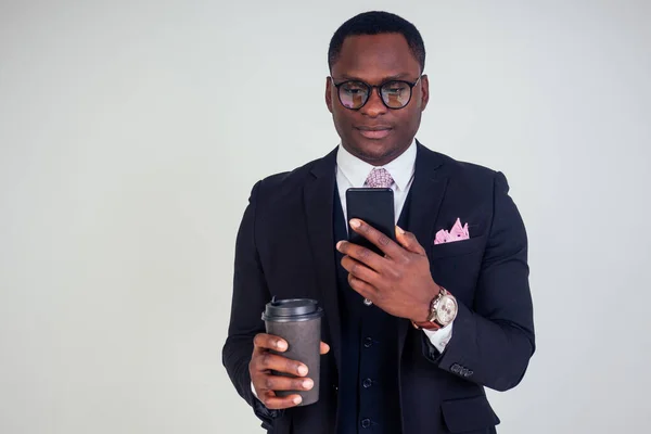 Handsome young African man in formal black suit holds a plastic paper cup with hot coffee cocoa tea to go and smartphone. businessman on coffee break after meeting on white background in studio
