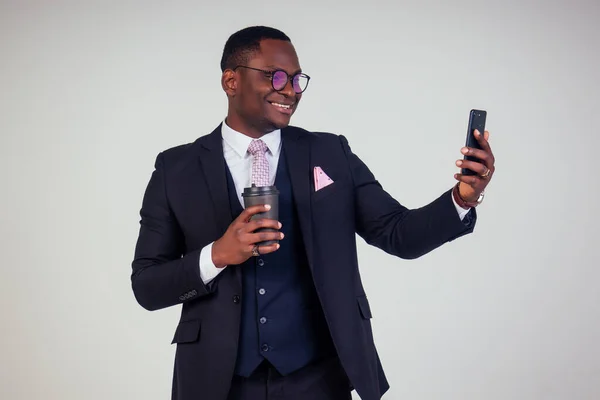 Handsome young African man in formal black suit holds a plastic paper cup with hot coffee cocoa tea to go and make selfie on smartphone. businessman on coffee break on white background in studio