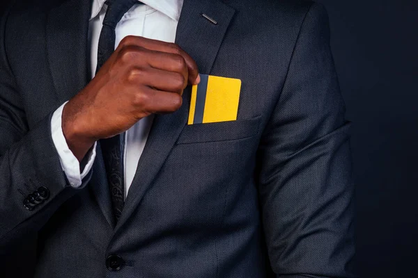 Joven exitoso hombre de negocios en un elegante traje clásico negro y en gafas frías con una tarjeta de crédito de plástico amarillo en el estudio sobre un fondo oscuro. concepto de compra — Foto de Stock