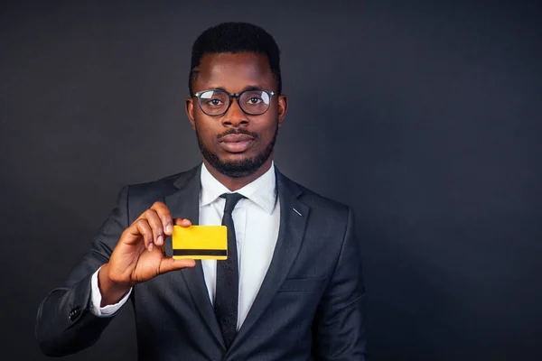 young successful businessman credit broker in a stylish black classic suit and in cool glasses holding a yellow plastic credit card in the studio on a dark background. shopping concept
