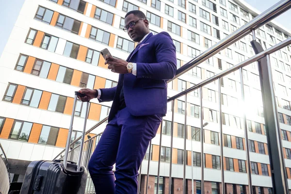 İyi giyimli ve başarılı bir Afrikalı Amerikalı iş adamı havaalanına giderken tekerlekli bavuluyla telefon ekranına bakıyor.