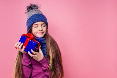 Mutlu, küçük, sarı kıvırcık saçlı, örgü ören gri şapkalı ve eşarplı, hediye kutusu pembe arka plan stüdyosu olan kız. Yeni yıl hediyesi, bir kadın çocuğun dilek kopyalama alanı.