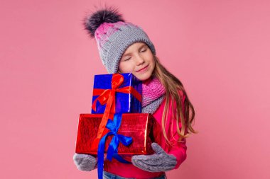 Pembe şapkalı, atkılı ve eldivenli, hediye kutusu pembe arka plan stüdyosu olan mutlu, kıvırcık saçlı kız. Yeni yıl hediyesi, bir kadın çocuğun dilek kopyalama alanı.