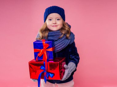 Pembe şapkalı, atkılı ve eldivenli sarı kıvırcık saçlı küçük bir kız hediye kutusu pembe arka plan stüdyosu. Yeni yıl hediyesi, bir kadın çocuğun dilek kopyalama alanı.