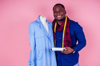American man working in a tailor workshop sews a blue coat on a pink background.young african fashion designer with mannequin at the tailoring studio clipart