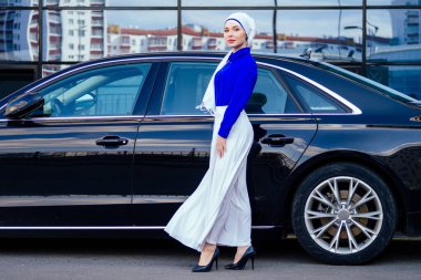 Arap Müslüman iş kadını tesettüre girmiş. Makyajlı. Lüks arabasının önünde duruyor. Kadın şık bir abaya gibi giyinmiş.