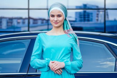 Arap Müslüman iş kadını tesettüre girmiş. Makyajlı. Lüks arabasının önünde duruyor. Kadın şık bir abaya gibi giyinmiş.