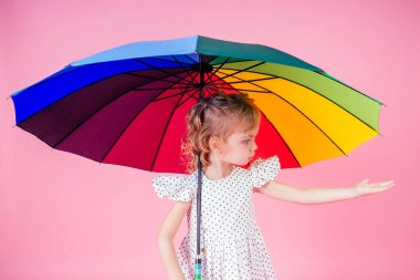 Mutlu ve güzel küçük sarışın kızın portresi. Bezelye rengi gökkuşağı şemsiyesi taşıyan beyaz elbiseli mavi gözlü kız. Stüdyoda bahar mevsimi pembe bir arka planda.