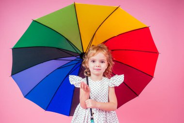 Mutlu ve güzel küçük sarışın kızın portresi. Bezelye rengi gökkuşağı şemsiyesi taşıyan beyaz elbiseli mavi gözlü kız. Stüdyoda bahar mevsimi pembe bir arka planda.